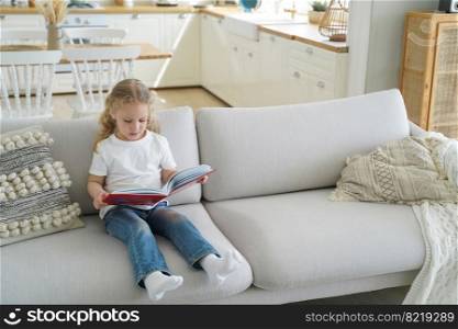 Small elementary girl reading fairy tale stories sitting on cozy sofa alone at home. Smart preschool child prodigy reads paper book, literature for children. Children’s education concept.. Small girl preschool child reads fairy tale stories holding paper book sitting on sofa alone at home