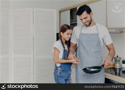 Small female child helps her father to prepare breakfast, fry eggs together, enjoy domestic atmosphhere, stand at kitchen, dad wears apron and holds frying pan. Family and cooking time concept