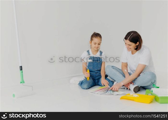 Small girl and her mother sit on floor, look attentively at color palette, choose best colour for painting room, use building tools, pose in spacious white room. Home renovation and design concept