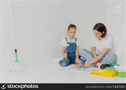 Small girl and her mother sit on floor, look attentively at color palette, choose best colour for painting room, use building tools, pose in spacious white room. Home renovation and design concept