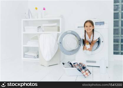 Small girl with appealing appearance, has fun and poses inside of washer, holds detergent, prepares for washing, basin with clothes for putting into washing machine on white floor. Laundry day at home