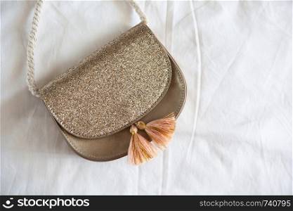 Small golden saddle handbag with tassels and rope handle lies on rumpled white sheet. Concept fees, outfitting, kid?s fashion, basic wardrobe. Copy space. Flat lay, top view