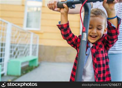 Small happy smiling boy standing on the electric kick push scooter holding the upper steering handle grip driven by his mother os aunt or other woman