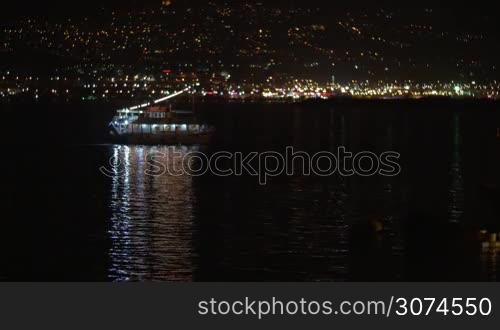 Small illuminated touristic boat sailing in the sea at night. Bright and colorful lights of coastal city in background
