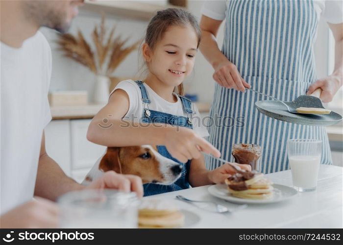 Small kid, her father and dog sit together at kitchen table, eat freshly prepared pancakes, mother in apron stands near holds pan. Family appetizing tasty desset at kitchen. Cooking, nutrition concept