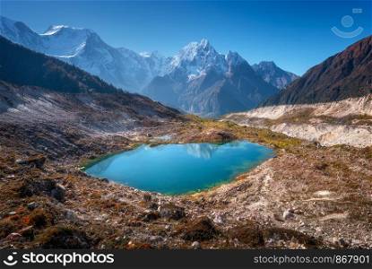 Small lake with blue water and sky reflection against beautiful snow covered mountains at sunny day in autumn. Landscape with lake, snowy rocks, hills, stones at sunrise. Himalayan mountains in Nepal