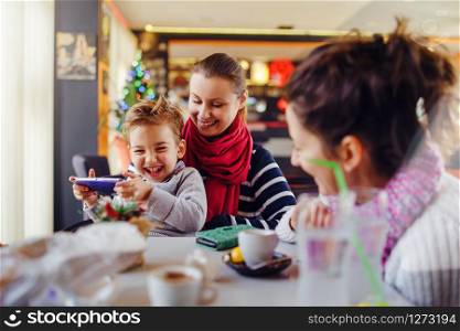 Small little boy caucasian child son sitting in the lap of his mother or aunt while female friend is looking at them teasing smiling holding smart mobile phone playing games or making a video call