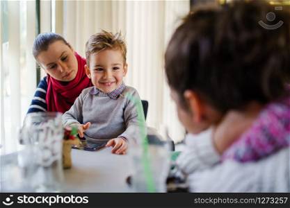 Small little boy caucasian child son sitting in the lap of his mother or aunt while female friend is looking at them teasing smiling holding smart mobile phone playing games making video call family