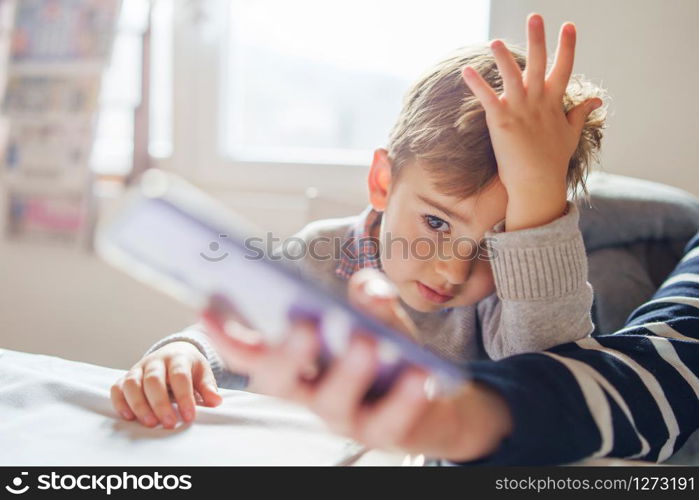 Small little boy sitting by the table at home or restaurant looking to the smart phone watching video or making video call