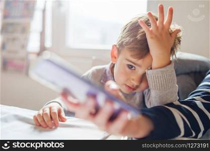 Small little boy sitting by the table at home or restaurant looking to the smart phone watching video or making video call