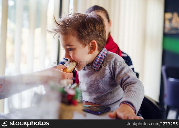 Small little boy three years old child son is sitting in the lap of his mother or aunt while somebody is feeding him having lunch at the table at cafe or restaurant