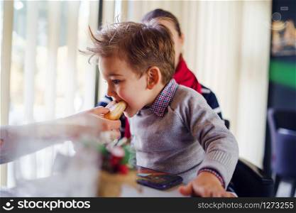Small little boy three years old child son is sitting in the lap of his mother or aunt while somebody is feeding him having lunch at the table at cafe or restaurant