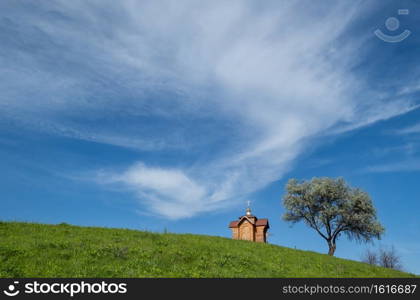 Small old wooden chapel on summer green grassy hill top, lonely willow tree and blue sky with cloud.