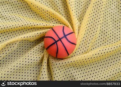 Small orange rubber basketball lies on a yellow sport jersey clothing fabric texture and background with many folds
