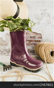 small plant planted purple wellington rubber boot with spool rope hat garden fork against weathered wall