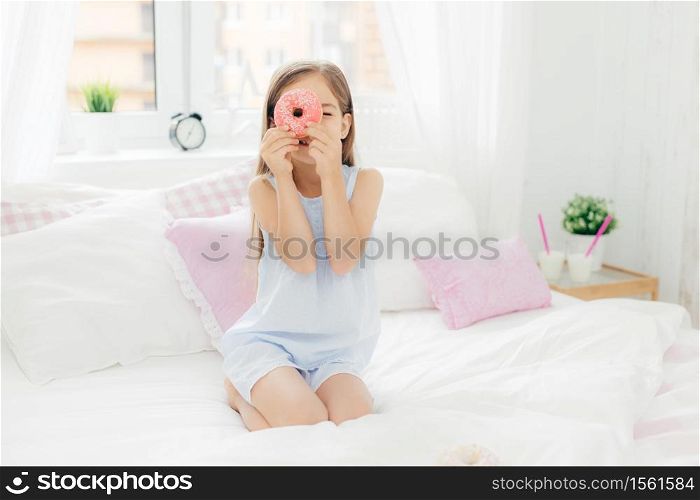 Small pretty female child holds tasty sweet doughnut, poses in bedroom on comfortabled bed, dressed in pyjamas, going to have breakfast, has joy and poses at camera. Chilhood and bedding concept