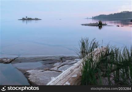 Small rocky islands with grass and driftwood among the clear waters of the lake at northern summer night. Reflection of the sky in mirror water. White Nights season in the Onega Lake - Karelia, Russia. Selective focus.