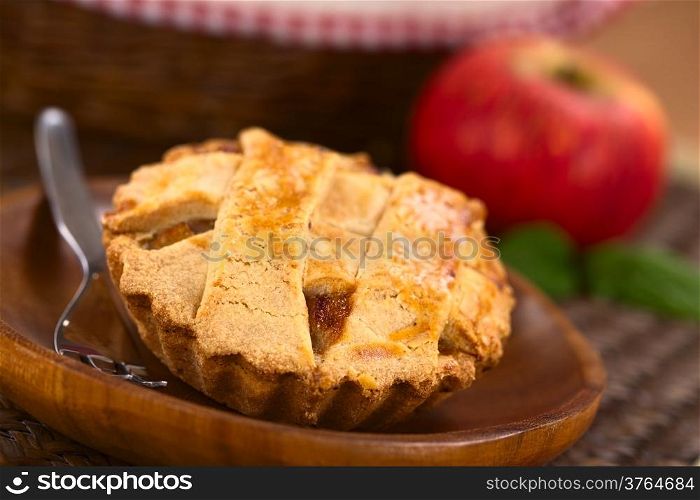 Small round apple pie with lattice crust on wooden plate with pastry fork, in the back an apple and a basket (Selective Focus, Focus one third into the pie)