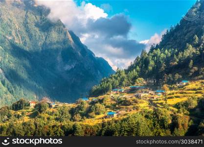 Small village on the hill lighted by a sunbeam against mountains and blue sky with clouds at sunset. Summer landscape with houses, fields with grass, gardens, green trees in Nepal. Rustic view. Travel. Small village on the hill lighted by a sunbeam against mountains