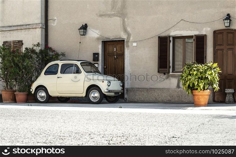 Small vintage italian car. Beige color old car in front of old house facade and flowers..