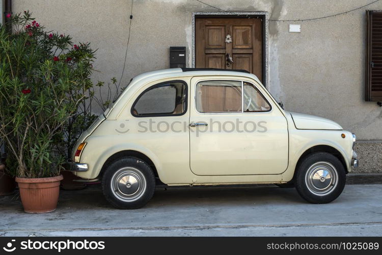 Small vintage italian car. Beige color old car in front of old house facade and flowers..