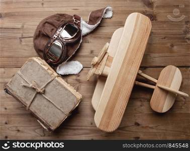 small wooden children's biplane plane on a wooden background next to a pilot helmet and glasses and a stack of old books. old wooden Airplane