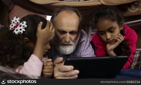 Smart adorable sisters explaining how to use internet via digital tablet to grandfather at home. Two little mixed race girls helping caucasian grandpa to use modern technology gadget as they lie together in cubby house. Dolly shot. Slow motion.