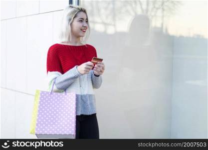 Smart and young consumer woman standing by a fashion store window display holding paper shopping bags and using a smartphone during a sunny day, outdoors.. young consumer woman standing by a fashion store window display holding paper shopping bags and using a smartphone