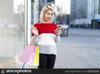 Smart and young consumer woman standing by a fashion store window display holding paper shopping bags and using a smartphone during a sunny day, outdoors.. young consumer woman standing by a fashion store window display holding paper shopping bags and using a smartphone