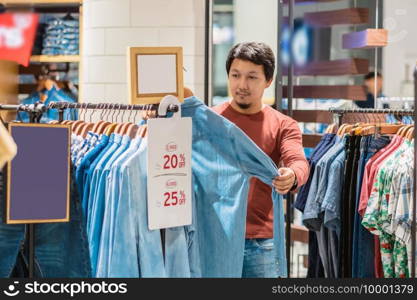 Smart asian man with beard choosing clothes in clothing store at shopping center, looking white jeans shirts , Fashion and Consumerism Concept.