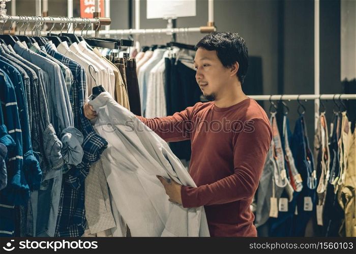 Smart asian man with beard choosing clothes in clothing store at shopping center, looking white color t-shirts , Fashion and Consumerism Concept.
