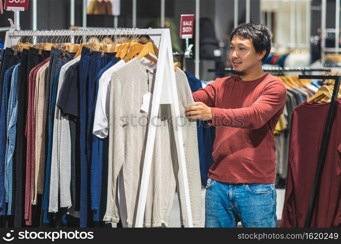 Smart asian man with beard choosing clothes in clothing store at shopping center, looking colorful t-sshirts , Fashion and Consumerism Concept.
