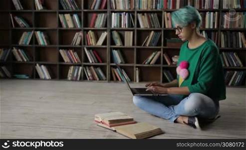 Smart college female student with blue hair e-learning with laptop and books. Young hipster woman in eyeglasses studying, reading educational material and typing on laptop pc while sitting on the floor in lotus posture over bookshelves background.