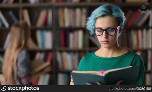 Smart confident blue hair nerdy woman in trendy spectacles absorbed in reading a book in university library. Another student searching for books on the background. Portrait of concentrated college student studying hard preparing for exam in library.