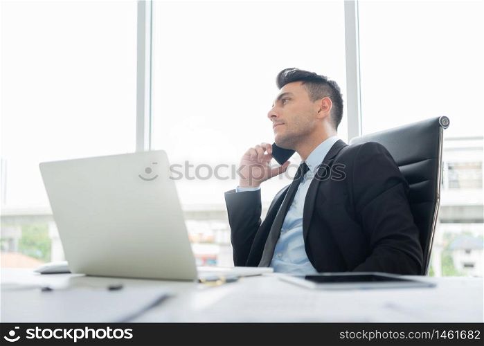 Smart Handsome Businessman in black suit is sitting using a mobile phone for negotiating business deals with partners and clients while working on a laptop computer at the workplace in the office.