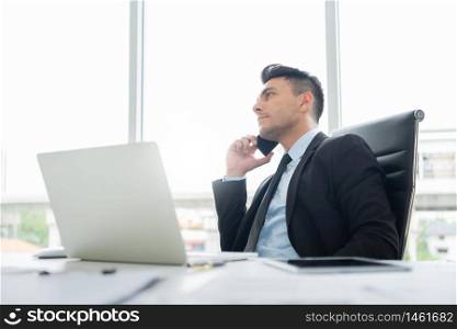 Smart Handsome Businessman in black suit is sitting using a mobile phone for negotiating business deals with partners and clients while working on a laptop computer at the workplace in the office.