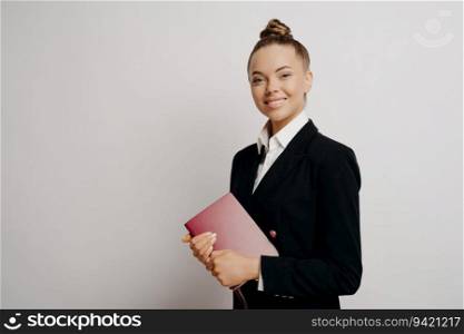 Smart, happy female business person holds red notebook, smiles at camera, posing isolated against light background.