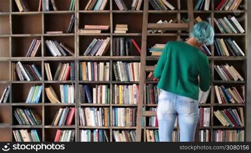 Smart hipster nerdy girl selecting new literature book from bookshelf in bookstore. Rear view. Young blue hair woman in trendy outfit standing and choosing book from shelf in library.