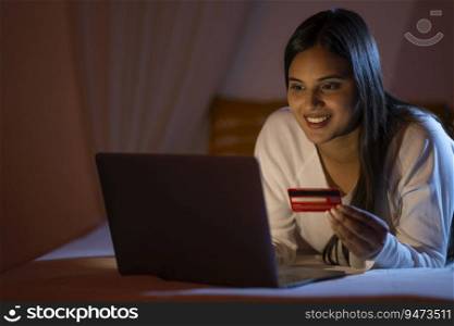 Smart Indian girl shopping online through laptop using debit card in bedroom