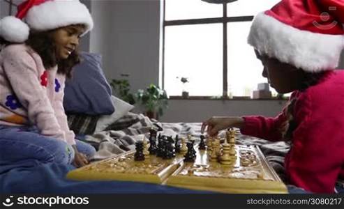 Smart small african american girls in santa hats playing chess on bed in domestic interior. Lovely serious sisters moving chess pieces on chessboard and capturing chess figures while enjoying time together on christmas at home. Dolly shot. Slo mo.