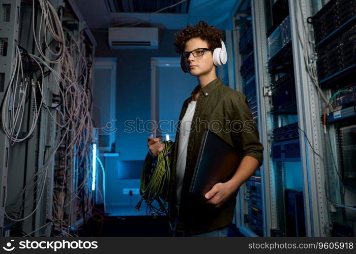 Smart teenager student having internship in modern data center. Male IT technician wearing headphones holding laptop computer carrying wires for solving problem with network connection. Smart teenager student having internship in modern data center