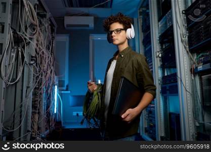 Smart teenager student having internship in modern data center. Male IT technician wearing headphones holding laptop computer carrying wires for solving problem with network connection. Smart teenager student having internship in modern data center
