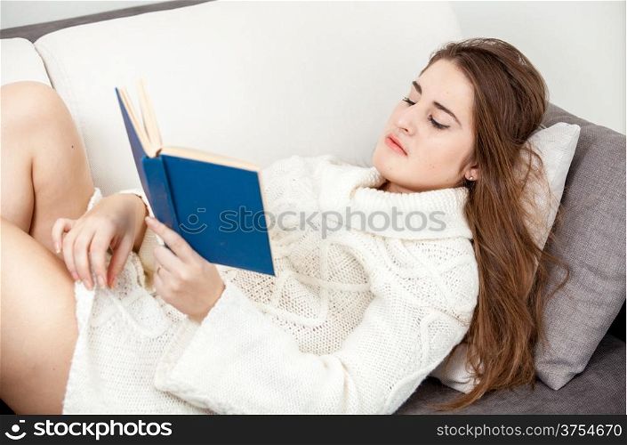 Smart woman in white sweater lying on sofa and reading book