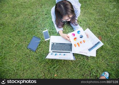 Smart woman working outdoor. Freelance Using laptop smart tablet and smart phone lay down on green grass field reading spreadsheet. work from home business concept.