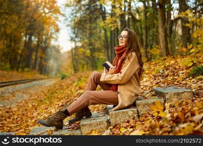 Smart young woman sitting on stairs reading book over autumn background. Golden leafage on ground. Young woman with book over autumn background