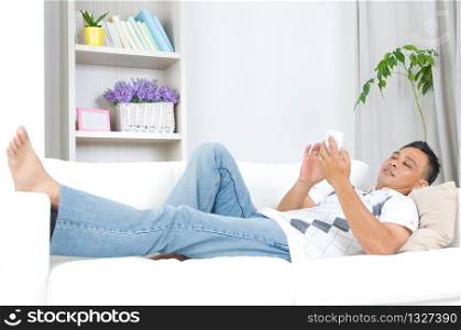 Smartphone mobile apps concept. Malay guy using smart phone. Handsome asian man relaxed and lying on sofa indoor.