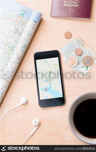 Smartphone with navigation map on screen, euro banknotes, paper map, passport, earphones on the wooden desk. Tourist essentials. Things related to travel