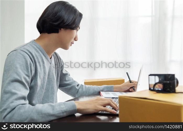 SME, Entrepreneur, Working concept. Business owner checking online orders with laptop computer at home. Young Asian man writing information from customers before preparing package products for delivery.
