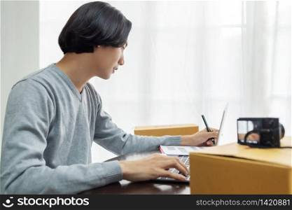SME, Entrepreneur, Working concept. Business owner checking online orders with laptop computer at home. Young Asian man writing information from customers before preparing package products for delivery.
