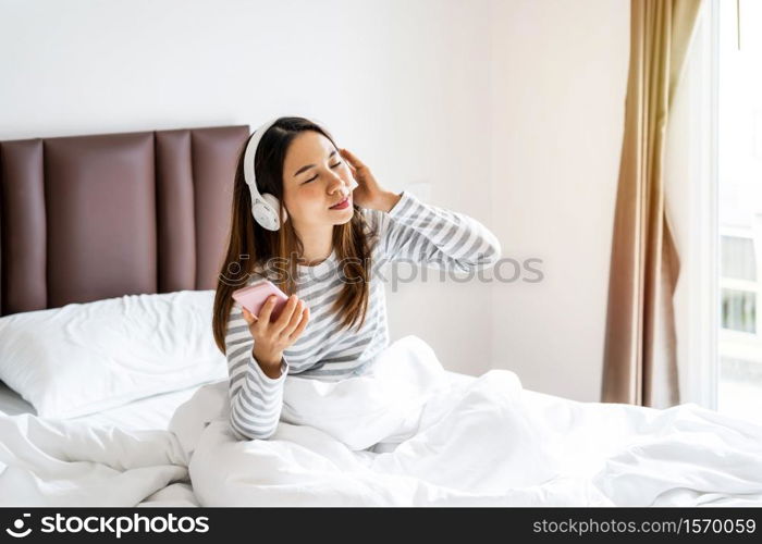 Smile happy young asian woman relaxing and using headphones to listen to music from smart phone in bedroom